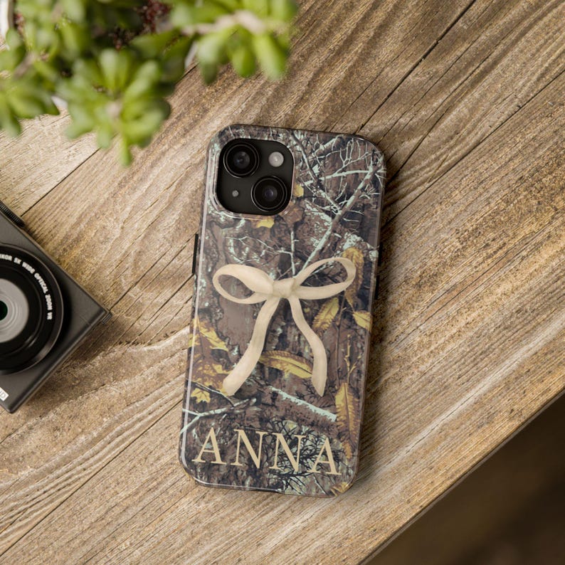May include: A phone case with a camouflage pattern, a cream-colored bow, and the name "ANNA" in a serif font. The case is on a wooden surface, with a camera and plant in the background.