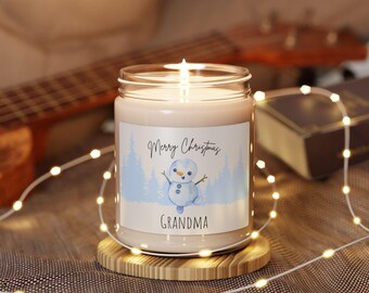 Pode incluir: Uma vela acesa em um frasco de vidro com um rótulo que diz "Merry Christmas Grandma" e apresenta uma ilustração de um boneco de neve. A vela está em uma base redonda de madeira. Luzes de fadas e uma guitarra são visíveis no fundo.