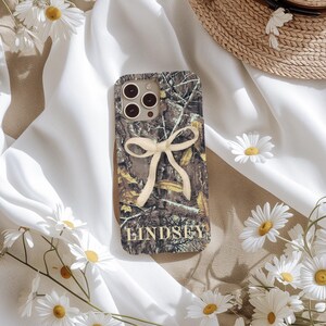May include: A camouflage-patterned phone case with a cream-colored bow and the name "Lindsey" in gold lettering. The case features a brown, green, and tan design, and is surrounded by white daisies and a straw hat.