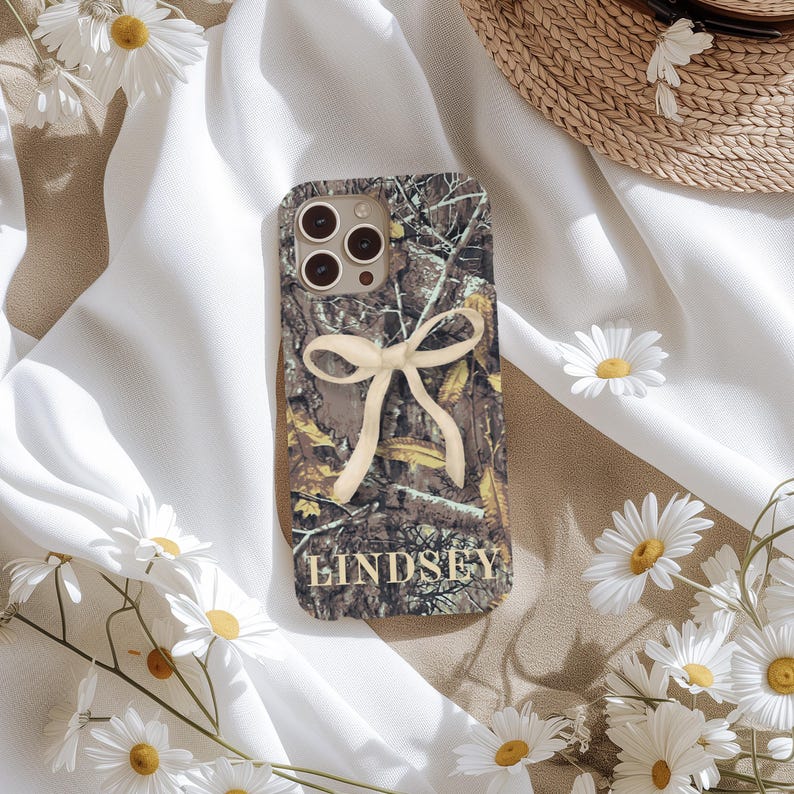 May include: A camouflage-patterned phone case with a cream-colored bow and the name "Lindsey" in gold lettering. The case features a brown, green, and tan design, and is surrounded by white daisies and a straw hat.