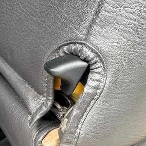 May include: Close-up of a black leather car seat with a black plastic lever. The seat has visible stitching and a textured surface. The lever is positioned near the seat's seam.