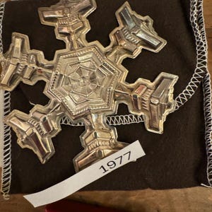 May include: A silver snowflake ornament with intricate details, displayed on a brown velvet pouch with a white label that reads "1977".