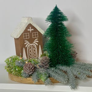 May include: A decorative Christmas display featuring a brown gingerbread house with white accents, a green bottle brush tree, pinecones, and faux greenery. The arrangement is set on a wooden base, creating a festive holiday scene.