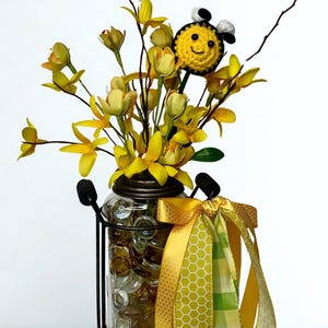 May include: A decorative jar with yellow flowers and a smiling bee topper. The jar is filled with clear stones and held in a black metal frame. A yellow and green ribbon is tied to the jar, adding a decorative touch.