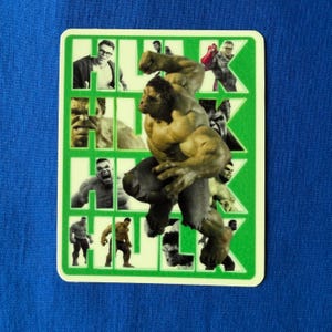 Può includere: Un adesivo rettangolare verde e bianco con più immagini di Hulk. L'immagine principale mostra Hulk in una posa dinamica, che rompe la griglia di immagini più piccole. Lo sfondo è blu.