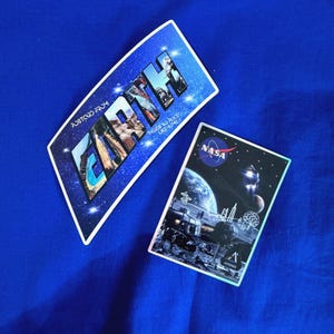 Puede incluir: Dos pegatinas sobre una superficie azul. Una dice "Postcard from Earth" con una escena espacial y la palabra "Earth". La otra tiene el logo de la NASA y una escena espacial con la palabra "Artemis".