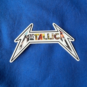 May include: A white and black sticker in the shape of a lightning bolt with the word "METALLICA" in colorful letters. The sticker is on a blue background.