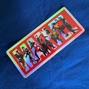 以下が含まれることがあります： 赤い背景に白い文字で「MARVEL」と書かれた長方形のマーベルステッカー。文字は切り抜かれており、さまざまなマーベルキャラクターの画像が表示されています。ステッカーにはホログラフィックで虹色のボーダーがあります。