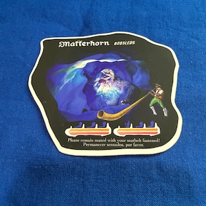 May include: A black, irregularly shaped card with a Matterhorn Bobsleds graphic. The image depicts a yeti in a cave, a horn player, and seating silhouettes. Text reads, "Please remain seated with your seatbelt fastened!"