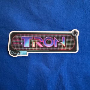 Könnte beinhalten: Ein rechteckiger Aufkleber mit dem Wort "TRON" in einem Farbverlauf aus Blau, Lila und Rosa. Der Text befindet sich vor einem schwarzen Hintergrund mit einem Rastermuster. Der Aufkleber hat einen weißen Rand und einen blauen Akzent auf der linken Seite.