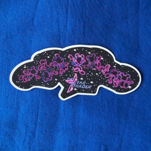 May include: A die-cut sticker with a black background, featuring a constellation of cartoon characters in pink and purple neon outlines. The sticker has a white border and the text "THE STAR TRADER" in neon pink and blue.
