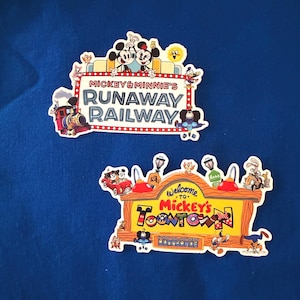 May include: Two die-cut stickers featuring Disney theme park signage. The top sticker displays Mickey & Minnie's Runaway Railway with a train and characters. The bottom sticker shows a Welcome to Mickey's Toontown sign with various characters.