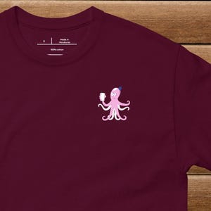 以下が含まれることがあります： 濃い赤紫色のTシャツに、コーヒーカップを持ったピンクのタコが描かれています。タコは頭に青い王冠をかぶっています。Tシャツには「ホンジュラス製」と「綿100％」のタグが付いています。