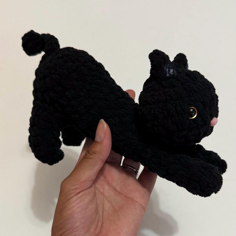 May include: A handmade black crochet cat toy with a pink nose and golden eyes. The cat is in a stretched-out pose, with its tail curled up. The toy is made of soft yarn.