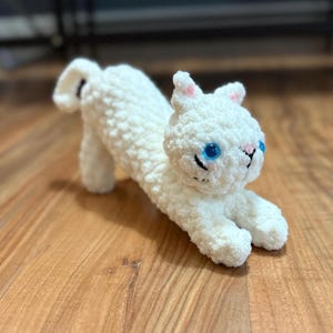 May include: A handmade white crochet cat toy with blue eyes, pink inner ears, and a black stitched mouth and nose. The cat is in a playful, stretched-out pose, showcasing its fluffy texture. The toy is placed on a wooden surface.