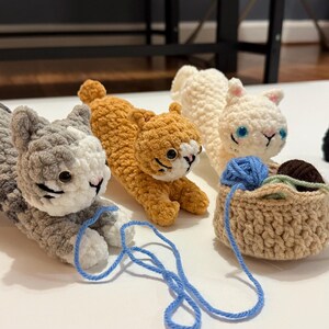 May include: A collection of handmade crochet cat figurines in various colors, including gray, orange, white, and black. Each cat has detailed features and is posed in different positions. A small crochet basket holds yarn.