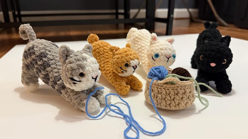 May include: A collection of handmade crochet cat figurines in various colors, including gray, orange, white, and black. Each cat has detailed features and is posed in different positions. A small crochet basket holds yarn.