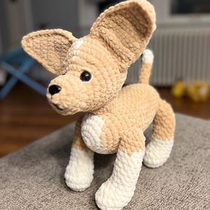 May include: A crocheted stuffed dog toy with brown and white fur. The dog is standing on a gray surface.
