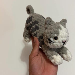 May include: A handmade crochet cat in shades of gray and white. The cat has black whiskers, a pink nose, and black eyes. The plush toy is designed to look like a cat in a playful pose.