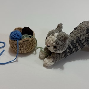 May include: A crocheted gray and white cat toy stretches towards a small basket filled with blue, green, and brown yarn. A length of blue yarn extends from the basket, suggesting play. The cat has white paws and a white face.