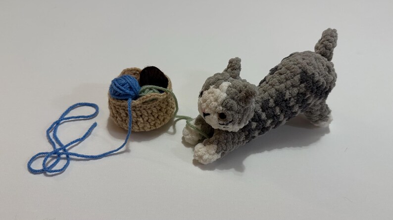 May include: A crocheted gray and white cat toy stretches towards a small basket filled with blue, green, and brown yarn. A length of blue yarn extends from the basket, suggesting play. The cat has white paws and a white face.