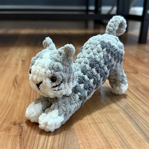May include: A handmade crochet cat toy in a stretching pose. The cat is primarily gray with white and darker gray patches. It has black whiskers, gold-colored eyes, and a pink nose. The toy is made of soft, plush yarn.