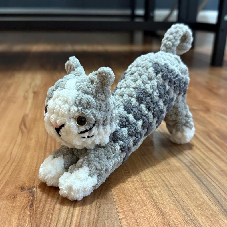 May include: A handmade crochet cat toy in a stretching pose. The cat is primarily gray with white and darker gray patches. It has black whiskers, gold-colored eyes, and a pink nose. The toy is made of soft, plush yarn.