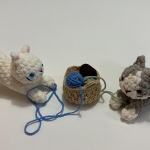 May include: Crocheted cat toys in white and gray, with blue yarn and a small basket. The white cat is playing with blue yarn, while the gray cat is playing with green yarn. The basket contains brown and green yarn. A charming cat-themed item.