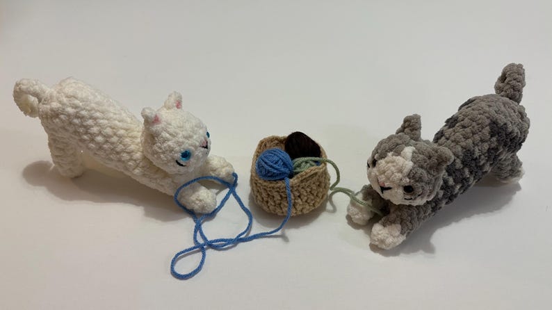 May include: Crocheted cat toys in white and gray, with blue yarn and a small basket. The white cat is playing with blue yarn, while the gray cat is playing with green yarn. The basket contains brown and green yarn. A charming cat-themed item.