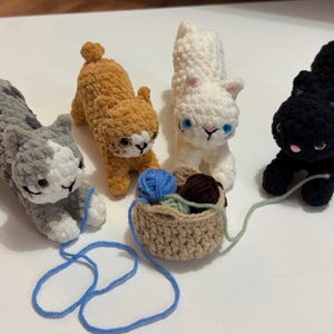 May include: Four crocheted cat figurines in gray, orange, white, and black. Each cat has a unique color pattern and is posed in a playful stance. A small basket holds yarn, with a blue strand extending towards a cat.
