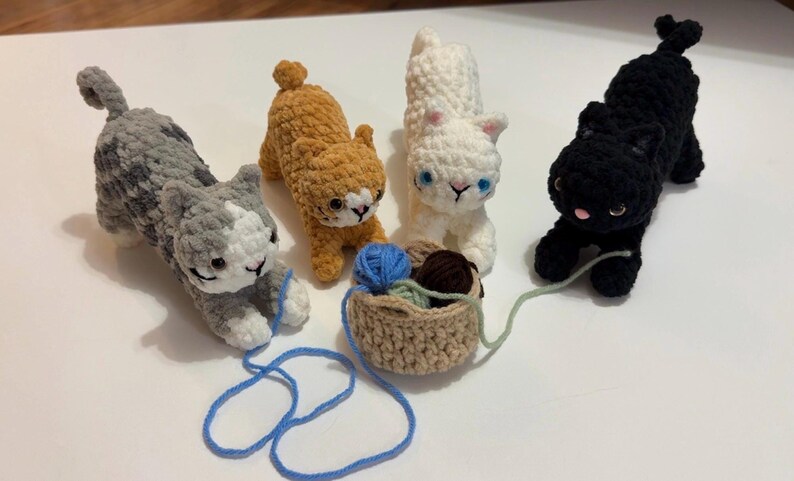 May include: Four crocheted cat figurines in gray, orange, white, and black. Each cat has a unique color pattern and is posed in a playful stance. A small basket holds yarn, with a blue strand extending towards a cat.