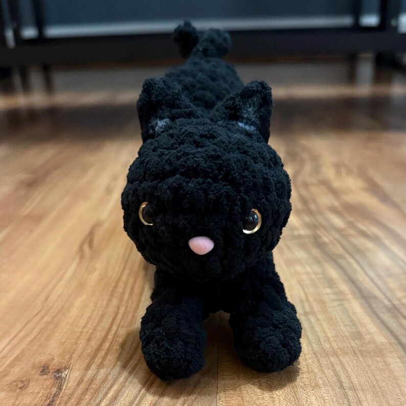 May include: A handmade black crochet cat plush toy. The cat has golden eyes, a pink nose, and small ears. The plush is lying down on a wooden surface. The cat's body is made of a soft, textured yarn.