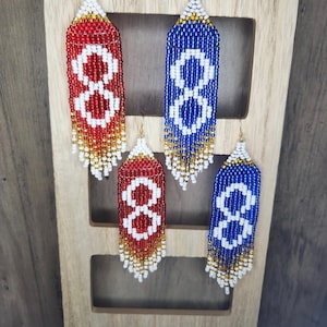 May include: Four pairs of beaded earrings with a fringe design. The earrings are red, white and blue with gold accents. The earrings are displayed on a wooden ladder.