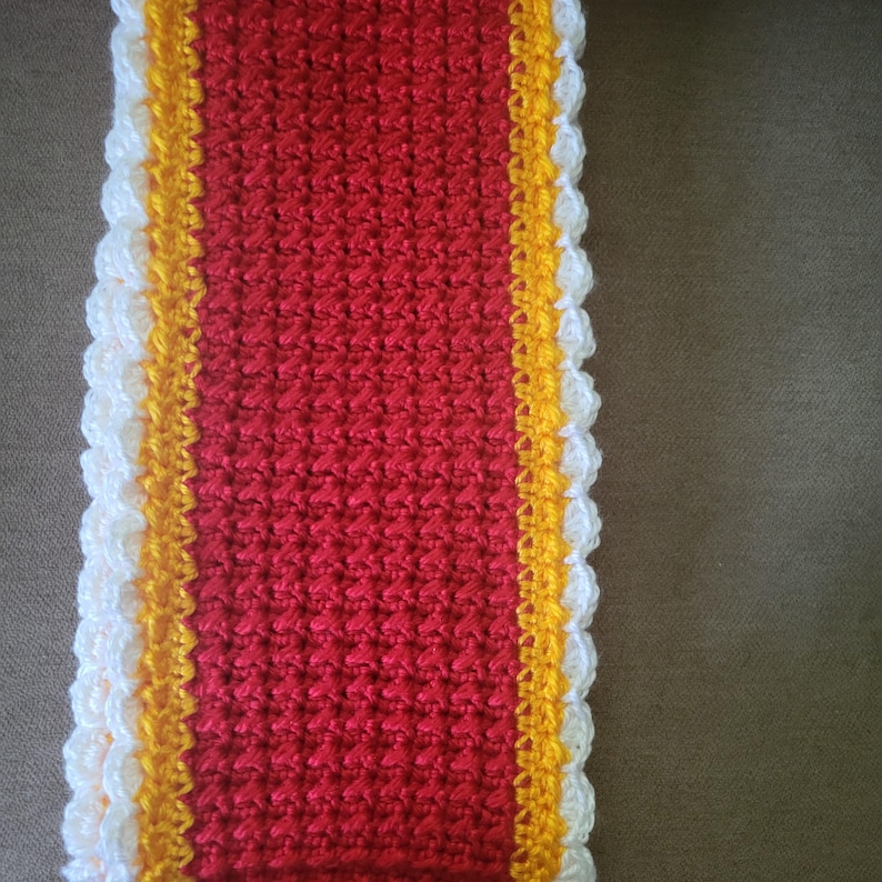 May include: A rectangular, hand-crocheted item with a red center, bordered by a yellow stripe and a white, scalloped edge. The textured stitchwork is visible. This item could be a scarf, table runner, or decorative piece.