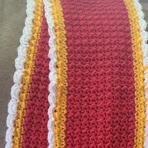 May include: A handmade crochet scarf in a vibrant red hue, accented with bands of yellow and white. The edges feature a scalloped trim, adding a decorative touch to this cozy accessory.