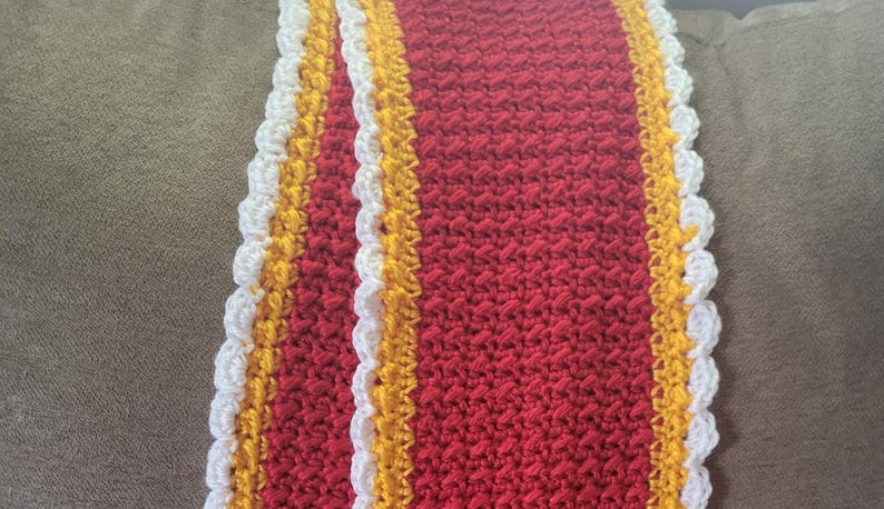 May include: A handmade crochet scarf in a vibrant red hue, accented with bands of yellow and white. The edges feature a scalloped trim, adding a decorative touch to this cozy accessory.