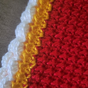 May include: Close-up of a crocheted item with a vibrant color scheme. The design features a scalloped edge in white, followed by a band of yellow, and a large field of red. The texture is clearly visible, showcasing the handmade quality.