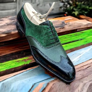 May include: Black leather oxford shoes with green suede accents and wingtip detailing. The shoes have a lace-up closure and are on a wooden surface.