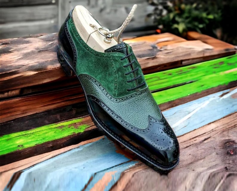 May include: Black leather oxford shoes with green suede accents and wingtip detailing. The shoes have a lace-up closure and are on a wooden surface.