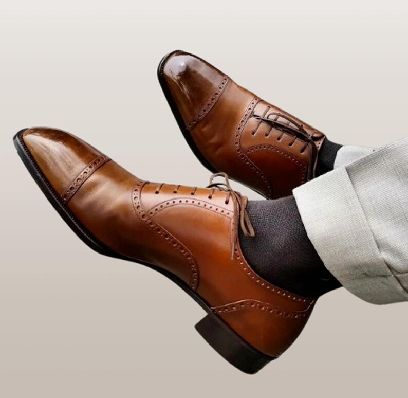K&ouml;nnte beinhalten: Ein Paar braune Lederschuhe mit Kappen und dekorativen Perforationen. Die Schuhe haben eine polierte Oberfl&auml;che und sind geschn&uuml;rt. Das Bild zeigt die Schuhe von unten, mit den Beinen und einer grauen Hose.