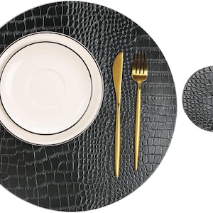 May include: A black placemat with a crocodile skin texture, a white plate with a black rim, and gold cutlery. A smaller matching coaster is also visible. The placemat is round and measures approximately 12 inches in diameter.
