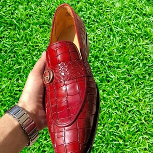 May include: A pair of red leather loafers with a crocodile pattern. The loafers have a gold buckle on the front.