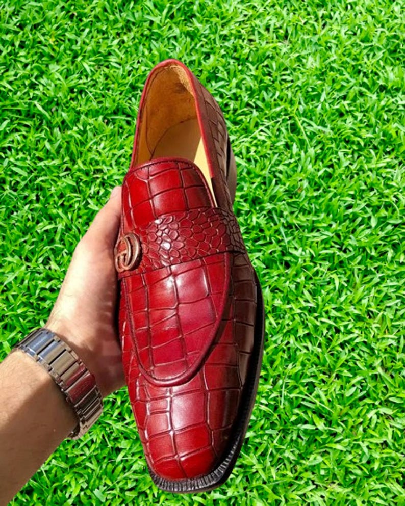 May include: A pair of red leather loafers with a crocodile pattern. The loafers have a gold buckle on the front.