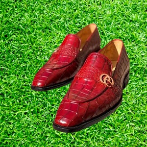 May include: A pair of red leather loafers with a crocodile pattern and a gold buckle. The loafers are on a green grassy surface.