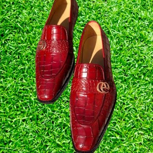 May include: A pair of red leather loafers with a crocodile pattern and gold accents. The loafers are on a green grassy surface.