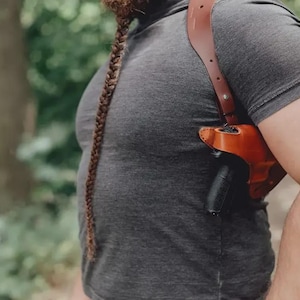 May include: A brown leather shoulder holster with a black handgun is worn over a gray t-shirt. The holster has a strap that goes over the shoulder and across the back. The background is blurred with green trees.