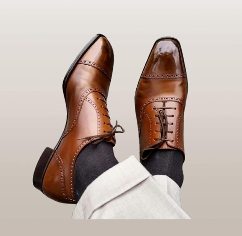 K&ouml;nnte beinhalten: Ein Paar braune Lederschuhe mit Kappe und Lyralochung. Die Schuhe haben eine polierte Oberfl&auml;che und sind mit braunen Schn&uuml;rsenkeln geschn&uuml;rt. Die Schuhe werden mit dunkelgrauen Socken und einer hellen Hose getragen.