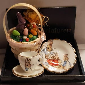 Op de afbeelding: Miniatuur Paas-thema set met een gevlochten mand gevuld met kleurrijke glazen eieren, wortelen en een paarse konijn. Inclusief een kopje, schotel en een schaal met geschulpte rand, allemaal versierd met konijnen illustraties. De set wordt gepresenteerd op een zwarte tray.