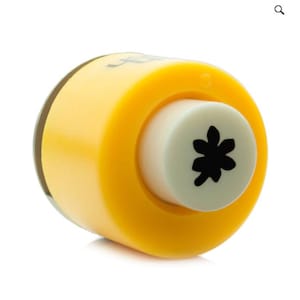 May include: A yellow and white flower-shaped paper punch. The punch has a yellow body with a clear plastic top and a white circular punch head. The punch head features a black flower design. This craft tool is used for paper crafting and scrapbooking.