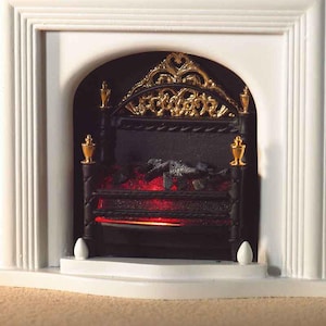 Könnte beinhalten: Ein Miniatur-Kamineinsatz mit einem schwarzen Metallgitter, goldenen Zierspitzen und einem verzierten goldenen Bogen. Im Inneren befinden sich dunkle, künstliche Kohlen, die von einem hellroten Licht beleuchtet werden, eingefasst in einen weißen Rahmen.
